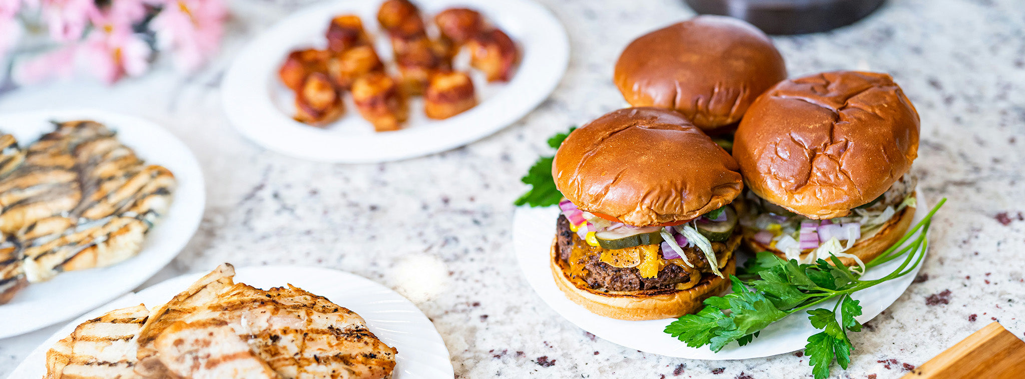 Burgers with buns on a plate, resting on a light marble table with plates of grilled chicken