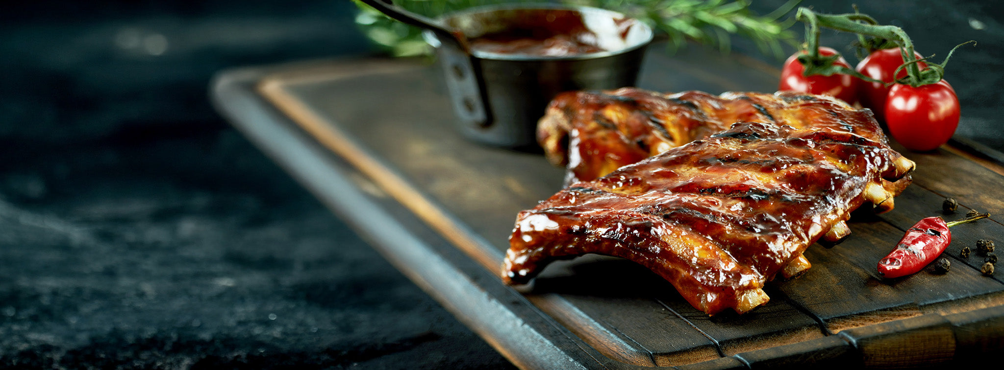 Saucy ribs on dark cutting board with tomatoes, herbs, and peppers